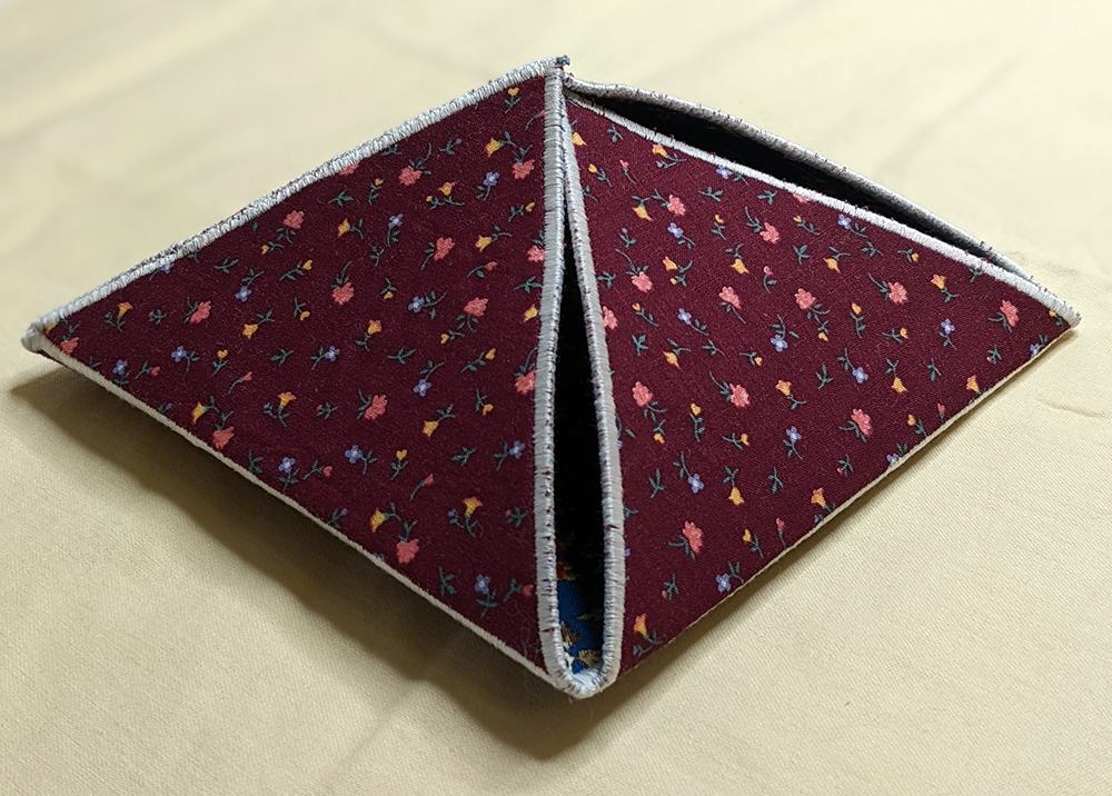 A red fabric box in the shape of a pyramid. This is a work in progress (WIP). The outside of the box is a dark red fabric with tiny pink, yellow, and blue print flowers. The open side is tucked into the top, keeping it mostly closed. Through one corner the inside fabric shows through, a blue floral print. The box is edged with white satin stitching all the way around each side, and around the 4-sided base.