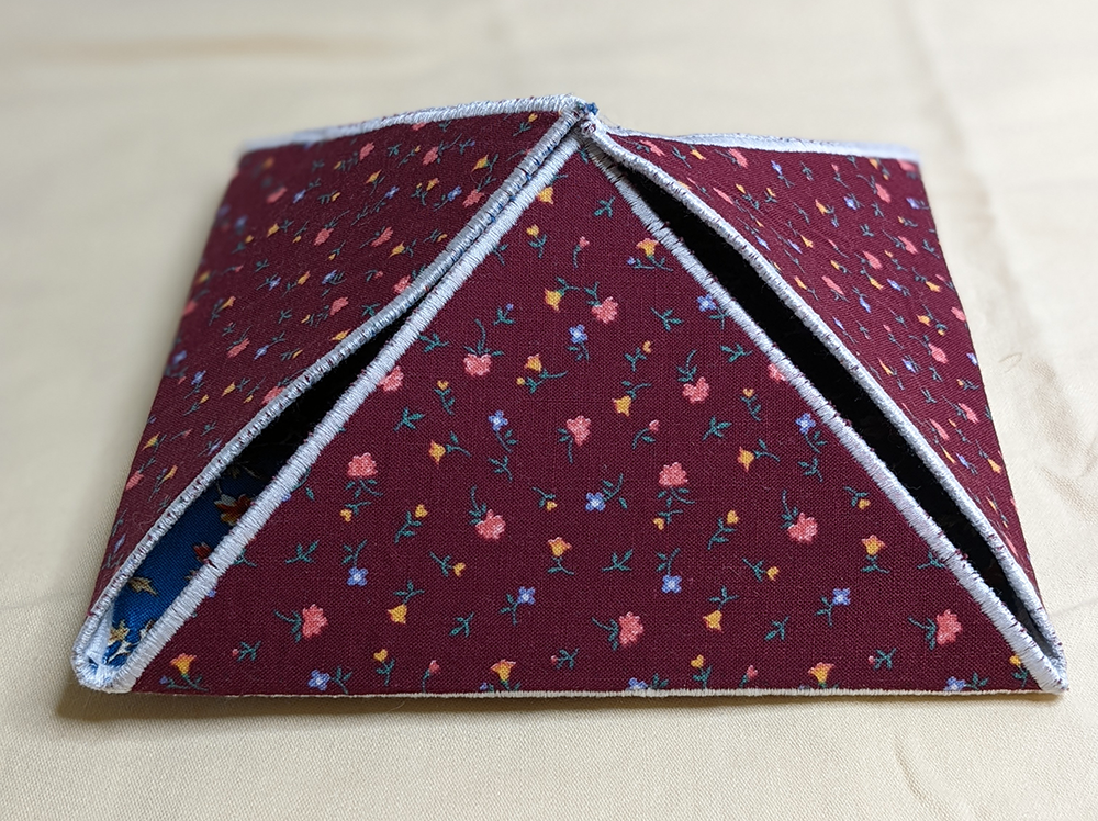 A red fabric box in the shape of a pyramid. This is a work in progress (WIP). The outside of the box is a dark red fabric with tiny pink, yellow, and blue print flowers. The open side is tucked into the top, keeping it mostly closed. Through one corner the inside fabric shows through, a blue floral print. The box is edged with white satin stitching all the way around each side, and around the 4-sided base.