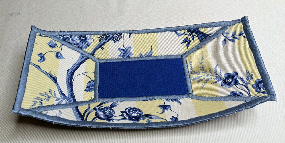 a small, rectangular fabric trinket tray, made from yellow, white, and blue print fabric. There is a blue rectangle of fabric centered in the tray, which is surrounded by light blue satin stitching. The satin stitching also extends from the corners of the blue rectangle out to the outer corners of the tray, and all around the edges of the tray.