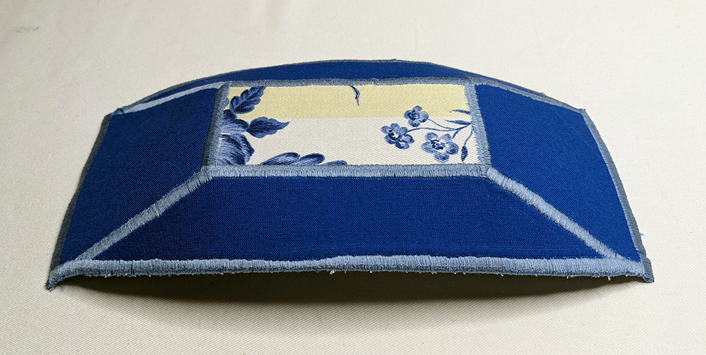 The outside of a small, rectangular fabric trinket tray, made from blue fabric. There is a rectangle of yellow, white, and blue print fabric centered in the tray, which is surrounded by light blue satin stitching. The satin stitching also extends from the corners of the blue rectangle out to the outer corners of the tray, and all around the edges of the tray.
